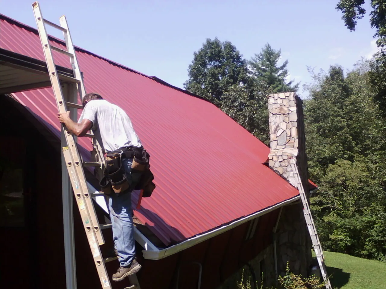 Roofer on a ladder beside a red metal roof and stone chimney — Soffit & Fascia Repair by Roofing Brothers in Juda